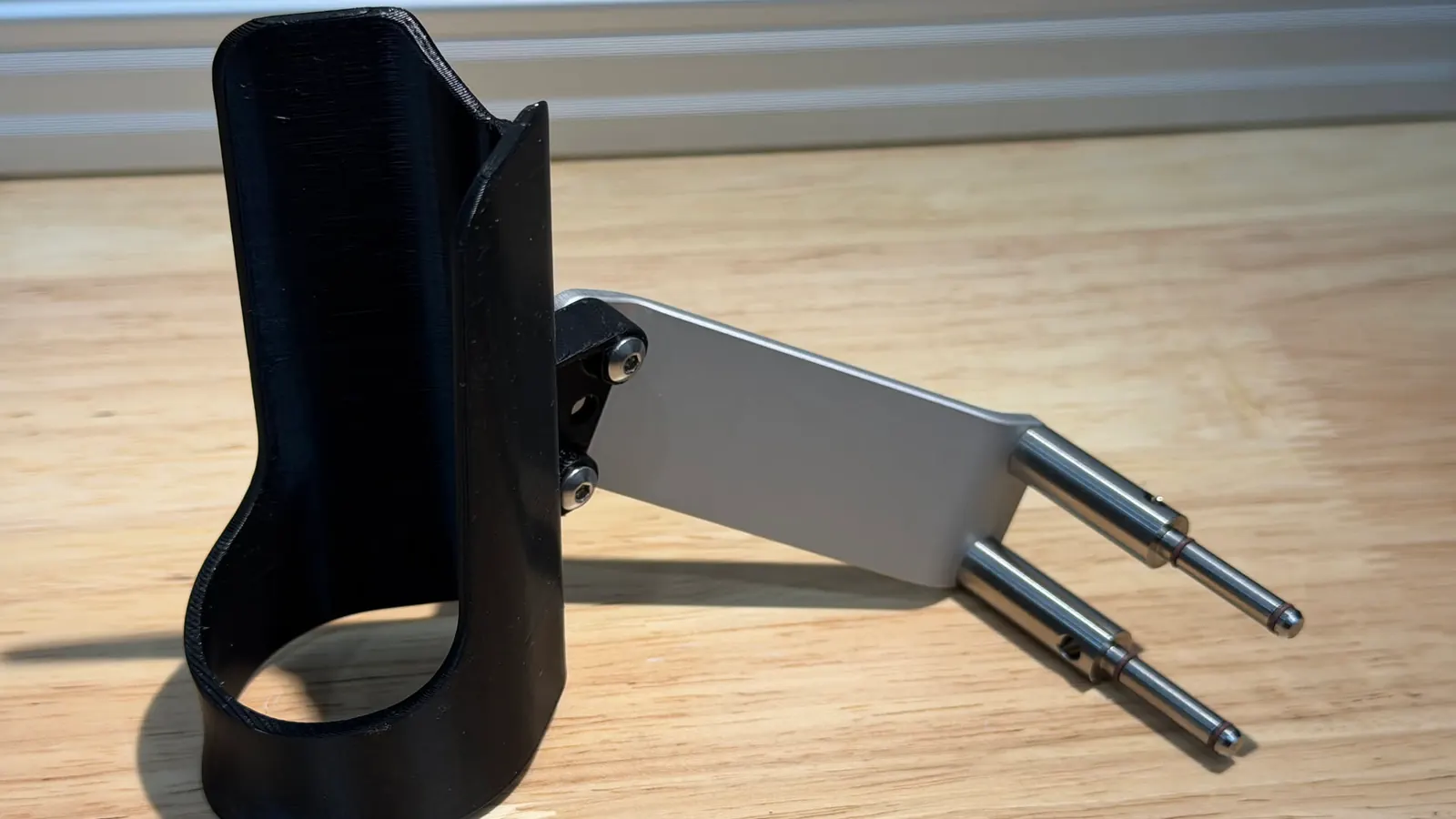 Black 3D-printed handpiece holder screwed to a silver aluminum mounting arm with two metal locating pins, on a wooden bench